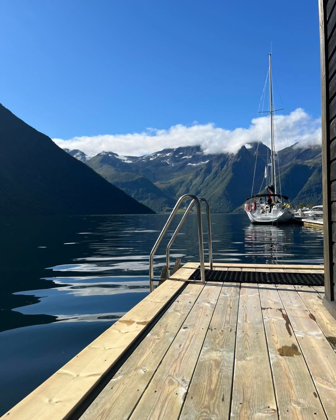 Badestigen og terrassen i sol, med ein seglbåt fortøydd ved sida og fjell med skyer bak.
