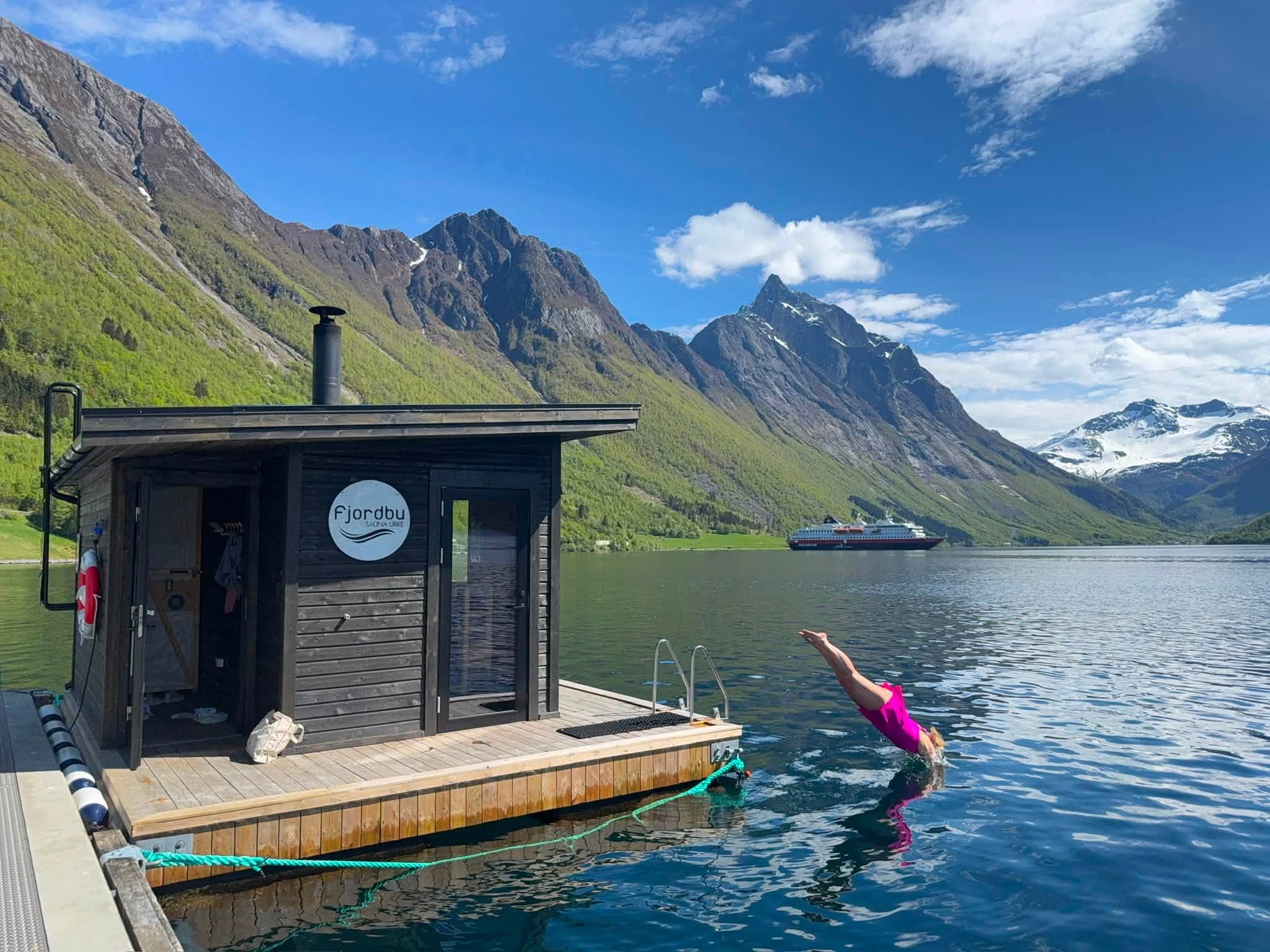 Badstua ved kaia med dørene opne, medan nokon stuper frå kanten ut i fjorden. Eit hurtigruteskip passerer under grøne fjellsider og snøkledde tindar.