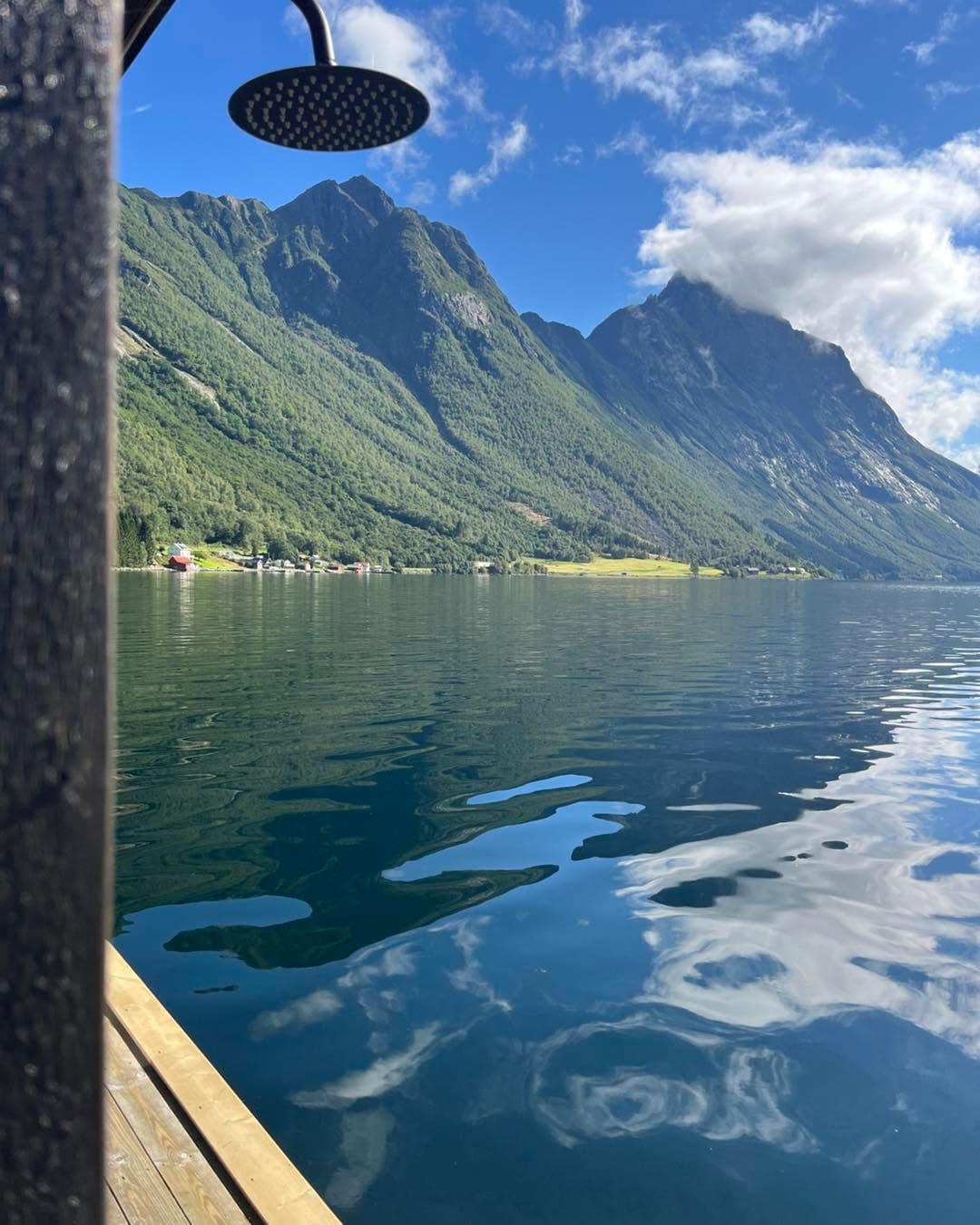 Utedusjen på kanten av flåten, med skyer spegla i blank fjord og grenda på andre sida.
